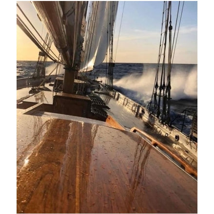 A close-up view from the deck of a tall ship sailing at sunset, with wet wooden hull, rigging, white sails, and ocean waves crashing alongside.