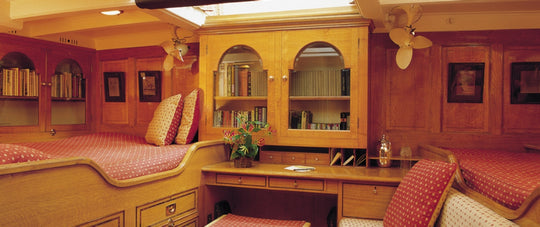 A warm wood-paneled interior room with built-in bookshelves, two beds with red patterned bedding, a desk, and ceiling fans.